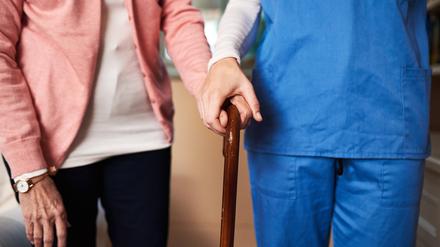 Eine blau gekleidete Pflegekraft unterstützt eine ältere Frau beim Gehen mit dem Gehstock. Prognosen zufolge wird sich die Personalnot in Pflegeberufen bis 2049 drastisch verschärfen.