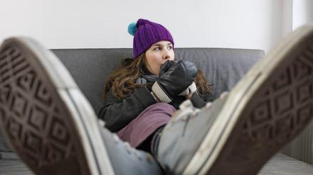 Junge Frau sitzt mit Mütze und Handschuhen auf dem Sofa: Heizkosten durch geringen Verbrauch sparen ist eine Variante. Manchmal gelingt es auch, die Kosten mit der Steuererklärung zurückzuholen.