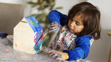Cute girl painting small wooden model house with paintbrush in living room.