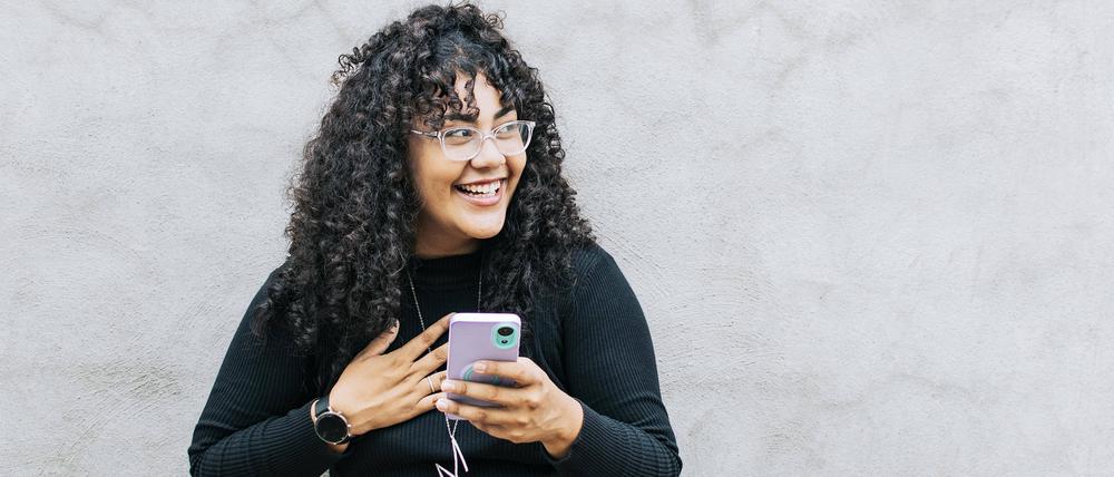 Eine junge Frau lachend mit ihrem Handy gegen eine Wand gelehnt.