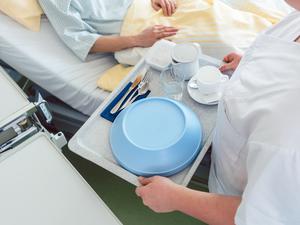 Eine Krankenpflegerin bringt ein Tablett mit Essen ans Bett einer Patientin.