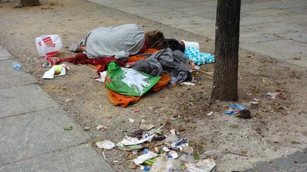 Ein Obdachloser mit wenig Habseligkeiten schläft am Straßenrand auf einer Decke zwischen Müll und Hundekot.