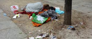 Ein Obdachloser mit wenig Habseligkeiten schläft am Straßenrand auf einer Decke zwischen Müll und Hundekot.