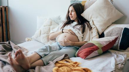 Eine junge schwangere Frau liegt in ihrem Bett und hält ihren Bauch.