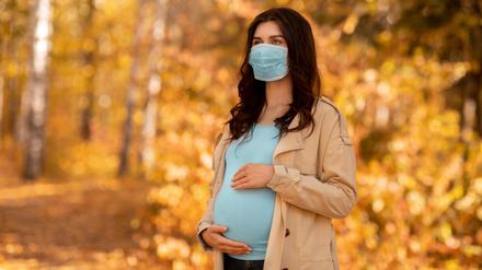 Young pregnant woman in medical mask on walk at autumn park during coronavirus epidemic. Expectant lady feeling lack of oxygen or morning sickness while wearing covid-19 protection outside